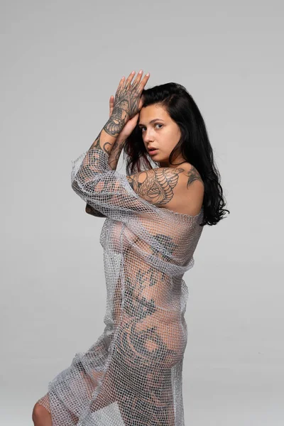 Stüdyoda çıplak poz veren genç ve güzel bir kız şeffaf bir elbisenin içinde duruyor.