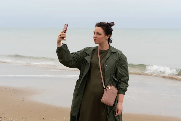 A girl stands with a phone near the sea in a raincoat