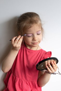 Küçük kız gözlerini boyar, kırmızı bir elbisenin içinde durur.