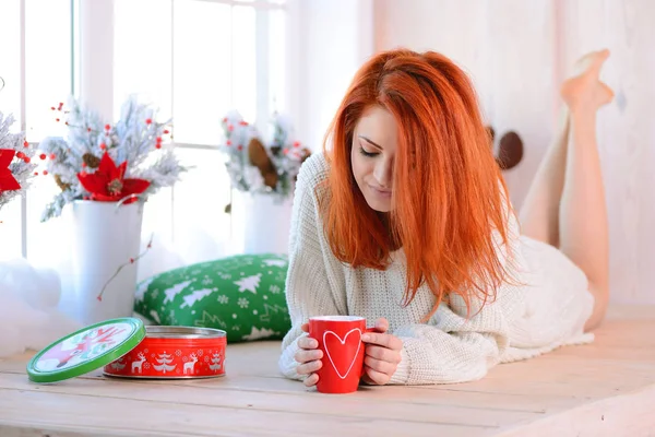 Schöne rothaarige Frau mit roter Tasse auf dem Boden liegend. das Konzept von Weihnachten. Weihnachten sonniger Morgen — Stockfoto