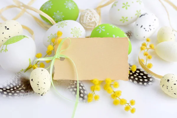 Huevos de Pascua y flores de mimosa sobre fondo blanco con tarjeta en blanco para saludar —  Fotos de Stock