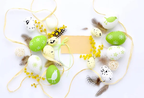 Huevos de Pascua y flores de mimosa sobre fondo blanco con tarjeta en blanco para saludar — Foto de Stock