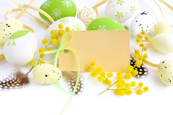 Huevos de Pascua y flores de mimosa sobre fondo blanco con tarjeta en blanco para saludar —  Fotos de Stock