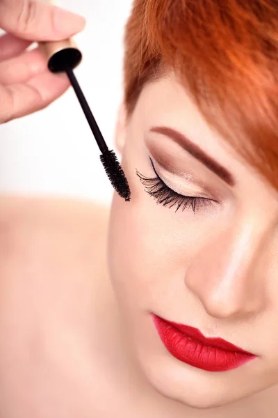 Maquillaje artista aplica rímel cepillo. Hermosa joven con el pelo rojo corto . — Foto de Stock