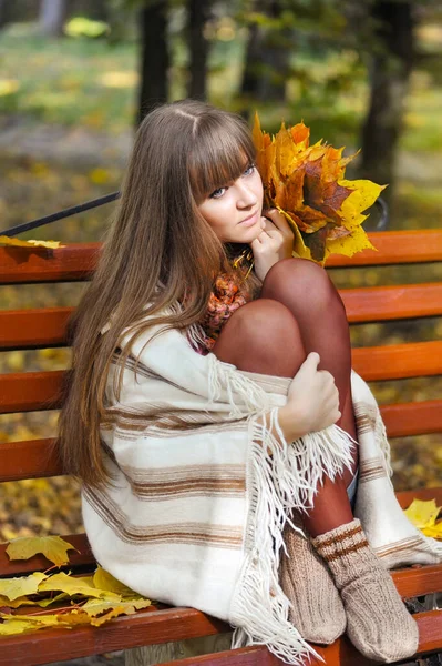 Schöne junge Frau sitzt auf einer Bank mit Herbstblättern — Stockfoto