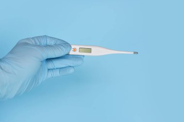 Eldivenler mavi arka planda dijital termometreyi tutuyor. Sağlık ve Tıp Konsepti.