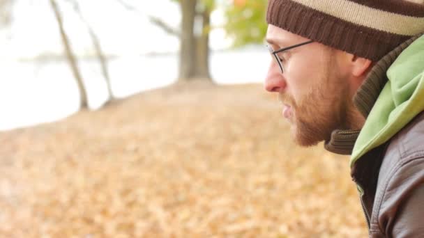 En skäggig man lampor en cigarett på en bänk i höst park. Tankeväckande allvarliga typ av symboliserar sorg — Stockvideo