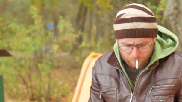 A bearded man in the autumn on a bench answers an incoming call on a mobile phone smoking a cigarette — Stock Video