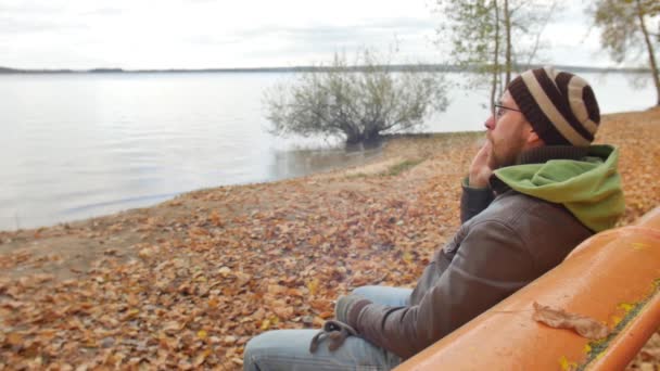 Бородач осенью на скамейке, разговаривая по мобильному телефону, соглашается и кивает головой, отрицая, что курил сигарету. — стоковое видео