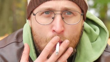 Sakallı bir adam odaya duman exhaling sigara bir bankta sonbaharda bir kazak ve deri ceketli