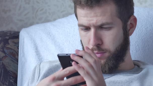 One white young brunette curly-bearded man uses the phone sitting in resle drinking tea or coffee — Stock Video