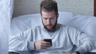 Bir beyaz genç esmer kıvırcık sakallı adam çay veya kahve içme bir sandalyede oturuyor telefon kullanır