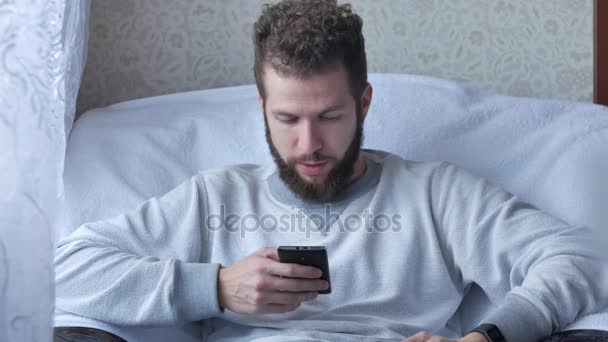 One white young brunette curly-bearded man uses the phone sitting in a chair drinking tea or coffee — Stock Video