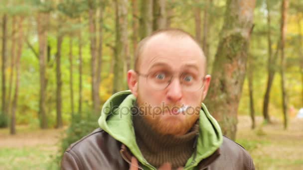 Een bebaarde man in een trui en lederen jas in de herfst schreeuwen en vloeken met een boos gezicht op de kijker roken van een sigaret uitademen van rook in de kamer in slow motion — Stockvideo
