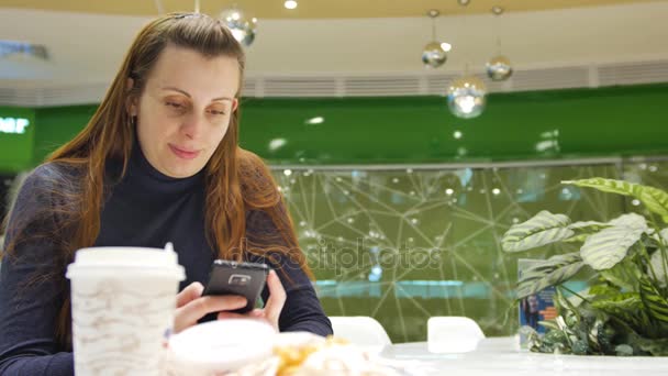 Een jonge blanke vrouw maakt gebruik van telefoon in een café in de supermarkt een openbare plaats. Een jonge blanke vrouw maakt gebruik van telefoon in een café in de supermarkt een openbare plaats. Hij wacht op zijn vriendin voor dranken — Stockvideo