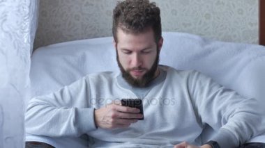 Bir beyaz genç esmer kıvırcık sakallı adam çay veya kahve içme bir sandalyede oturuyor telefon kullanır