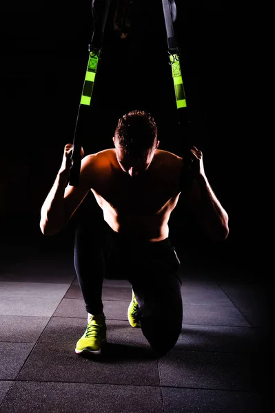 Ένα νεαρό όμορφος γενναία σγουρά γενειοφόρος μυώδης άνδρας με μούσι κάνει συνολική αντίσταση ασκήσεις γυμναστικής ιμάντες trx ασκηθείτε στο γυμναστήριο χαμηλών τόνων — Φωτογραφία Αρχείου