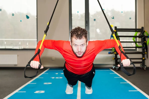 Um homem esportivo de cabelos encaracolados e barbudo branco que exercita o push-up de suspensão com alças de fitness trx no ginásio — Fotografia de Stock