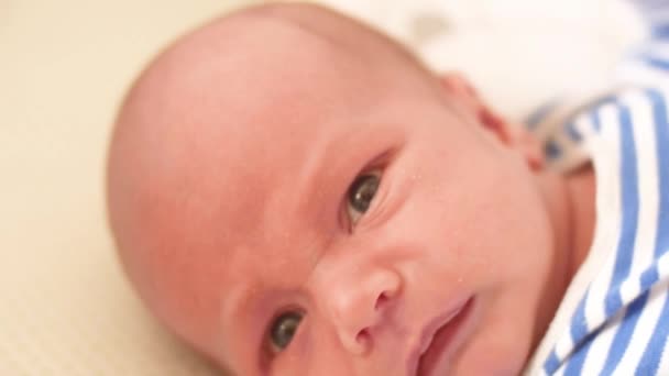 Un lindo bebé está mirando a la cámara y en una sábana blanca. El bebé mira a su alrededor y luego sonríe y ríe . — Vídeos de Stock