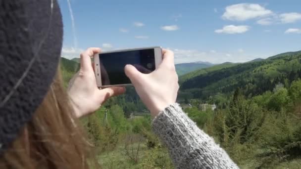 A woman is taking pictures against the background of big mountains and the green mountain river. on the phone. selfie or self-portrait on a smartphone. Enjoys adventure and travel concept — Stock Video