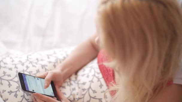 Sexy blond girl talking on mobile phone in bed, using touchscreen phone, browsing the Internet, chatting with someone. On the top hang a white canopy. Using mobile devices in everyday life — Stock Video