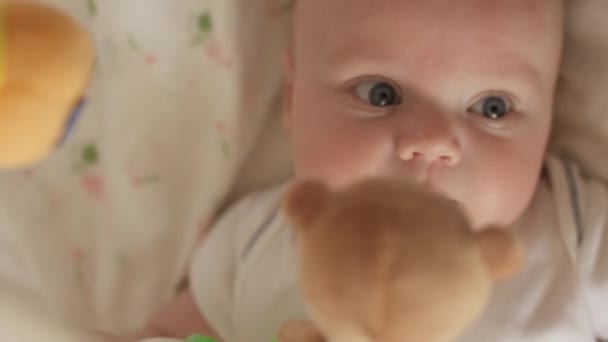 Vista superior de tiro de bebé lindo con chupete tumbado en la cuna y mirando girar juguetes oso taddy. El bebé mira a su alrededor y luego sonríe y ríe. Concepto de cuidado de los niños, amor paternal y carencia — Vídeos de Stock