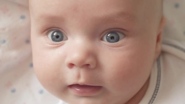 O olhar do bebê na câmera de perto tiro. Um bebé giro está a olhar para a câmara. O bebê olha em volta e depois sorri e ri. Conceito de cuidar de crianças, amor parental e — Vídeo de Stock