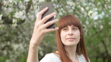 Bir kız bahçede selfie çekiyor. Çekici kızıl saçlı bir kadın kiraz bahçesinde cep telefonu kullanarak selfi yaparken gülümsüyor. Cihazları sağlıklı bir yaşam tarzı için kullanma kavramı.