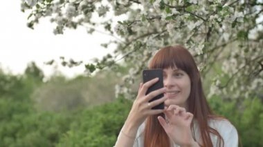 Kız bahçede selfie çekiyor. Çekici kızıl saçlı bir kadın kiraz bahçesinde cep telefonu kullanarak selfi yaparken gülümsüyor. Cihazları sağlıklı bir yaşam tarzı için kullanma kavramı.