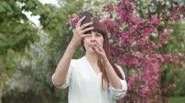 Kız bahçede selfie çekiyor. Çekici kızıl saçlı bir kadın kiraz bahçesinde cep telefonu kullanarak selfi yaparken gülümsüyor. Cihazları sağlıklı bir yaşam tarzı için kullanma kavramı.