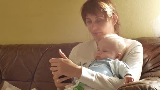 Καυκάσιος μεσήλικας ευτυχισμένη γυναίκα ψάχνει στο κινητό τηλέφωνο, ενώ το βρέφος γιο της παίζοντας σχετικά με τα χέρια της. Εσωτερικη πλάνα — Αρχείο Βίντεο