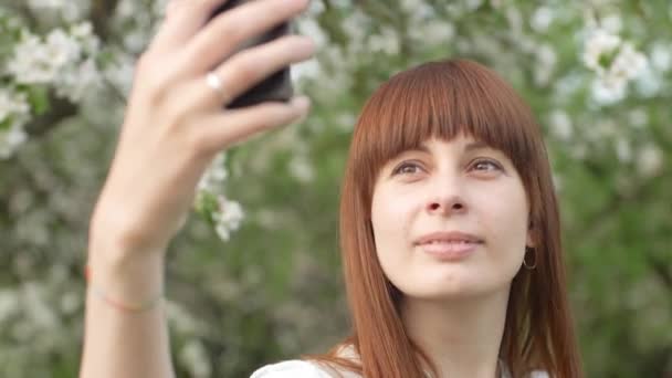 庭で女の子が自撮りをする。桜の果樹園で携帯電話を使って自撮りをしている魅力的な赤い髪の女性の笑顔。健康的なライフスタイルのためのガジェットの使用の概念. — ストック動画