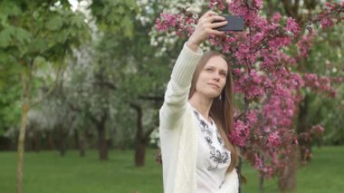 Kız bahçede selfie çekiyor. Çekici kızıl saçlı bir kadın kiraz bahçesinde cep telefonu kullanarak selfi yaparken gülümsüyor. Cihazları sağlıklı bir yaşam tarzı için kullanma kavramı.