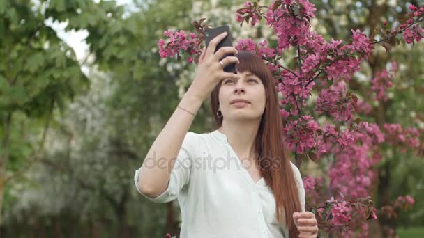少女は庭で自撮りをする。桜の果樹園で携帯電話を使って自撮りをしている魅力的な赤い髪の女性の笑顔。健康的なライフスタイルのためのガジェットの使用の概念. — ストック動画
