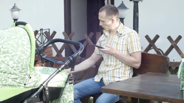 Un hombre con un niño en un cochecito se sienta en un banco, usa un teléfono y bebe café. — Vídeos de Stock