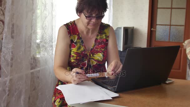 Oudere vrouw die op laptop, werkt het maken van notities op papier — Stockvideo