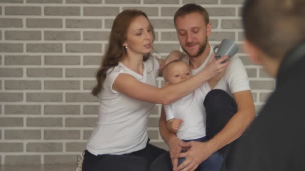 En lycklig familj deltar i en fotosession. Glada unga föräldrar med ett barn skrattar och poserar framför en fotograf — Stockvideo