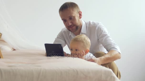 Un joven padre le muestra algo en una tableta a su pequeño hijo — Vídeo de stock