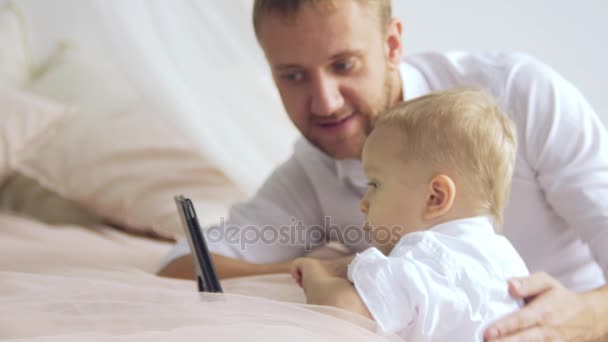 Молодий батько показує щось на планшеті своєму маленькому синові. Тато обіймає сина — стокове відео