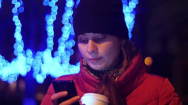 Una chica en la calle con una taza de café en sus manos está leyendo algo por teléfono y sonriendo, cha — Vídeos de Stock