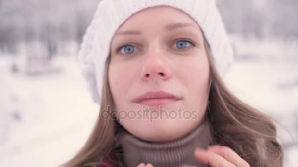 Una ragazza con bellissimi occhi azzurri guarda la fotocamera e sorride nella luminosa giornata invernale soleggiata. Parco innevato sullo sfondo — Video Stock