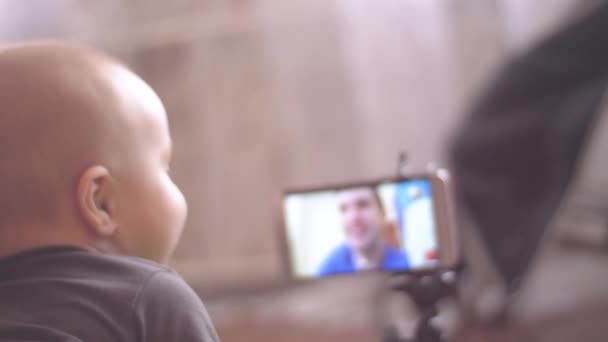 Babyjongen kijkt naar de Kaukasische papa op skype in een smartphone en het scherm raakt. 4 k binnen achteraanzicht close-up beelden. — Stockvideo