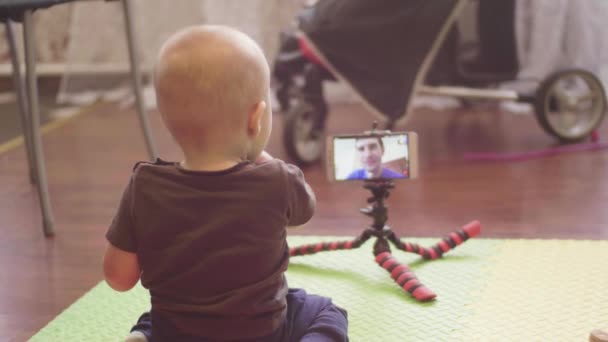 Small baby sitting on the floor communicates on video call in a smartphone with his caucasian father. Indoor 4K back view footage. — Stock Video