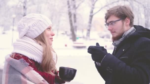 Hermosa mujer y hombre barbudo con gafas hablando y bebiendo cacao caliente de tazas en sus manos en el parque de invierno. Imágenes de primer plano de la vista lateral 4K . — Vídeos de Stock