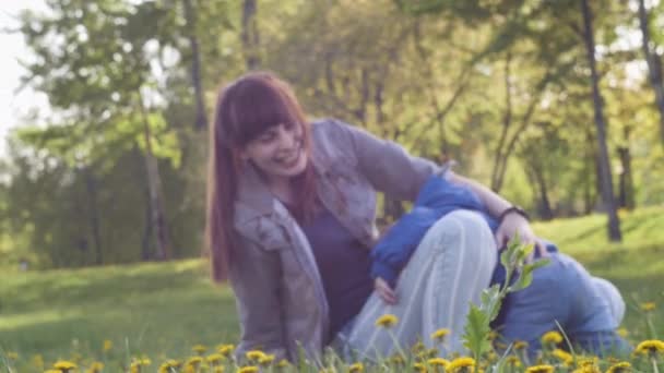 Ładna kobieta bawi się i bawi ze swoim synkiem ubranym w niebieską kurtkę na polanie mniszka lekarskiego w wiosennym, słonecznym parku miejskim z kwitnącymi drzewami na tle. 4k powolny film wideo. — Wideo stockowe