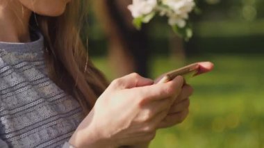 Güzel bir kadın, Spring City Park 'ta yürürken arkaplandaki elma ağacının çiçek açan dalında akıllı telefondan mesaj atıyor. Gün ışığında ve işaret fişeğinde dışarıda mafya telefonu kullanan bir kız. Yavaş çekim 4k video.