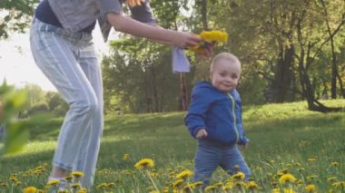 Genç bir kadın oğluna karahindiba çelengi takıyor ve oğluyla arka planda çiçek açan ağaçlarla bahar güneşli şehir parkında çimlerin üzerinde eğleniyor. 4k ağır çekim videosu.