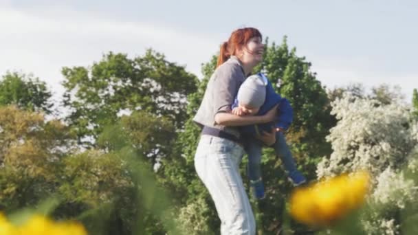 Mooie vrouw cirkelt baby boy gekleed in blauwe jas op haar handen en plezier met kind in het voorjaar zonnige stadspark met bloeiende bomen op de achtergrond. 4k slow motion video. — Stockvideo