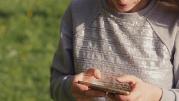 Tânăra femeie trimite mesaje text folosind smartphone-ul pe fundalul unui măr înflorit la mersul pe jos în parcul orașului de vară. Fata folosind telefonul moblie în aer liber. Slow motion 4K video . — Videoclip de stoc