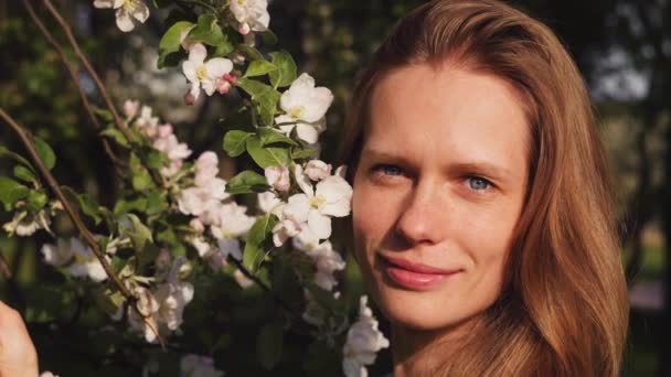 Porträt einer glücklichen schönen Frau, die an einem sonnigen Frühlingstag neben einem blühenden Apfelbaum steht, im Hintergrund der Stadtpark. blickt das Mädchen in die Kamera, lächelt und zwinkert. Zeitlupe 4k Video. — Stockvideo